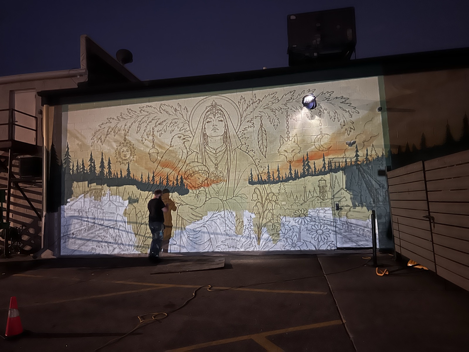 At night, the full line drawing projected onto the wall over the sky underpainting, an artist tracing it by hand