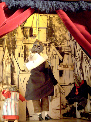 The marionette stage: a grey hunchback between two child puppets, against a pen-and-ink castle town drawn onto the boards.