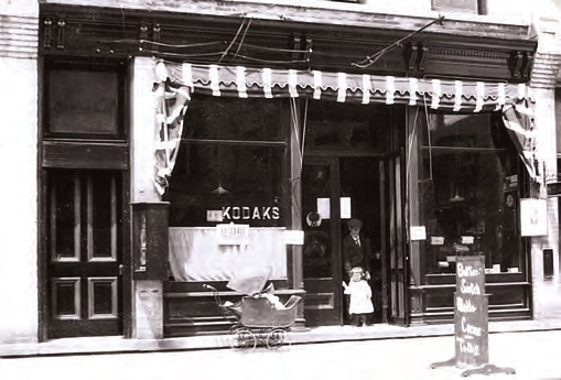 The Wilbur Building storefront in 1917 as Fluck's Drug Store — twin display windows, a striped awning, and KODAKS lettered on the glass.