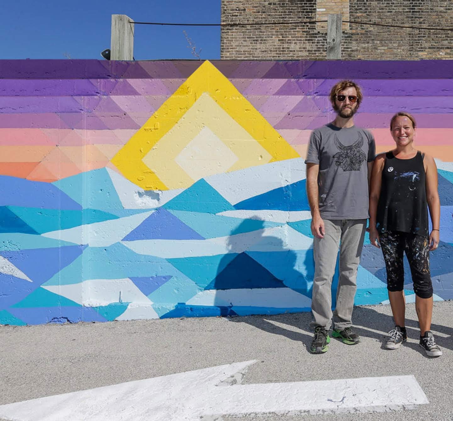 Don Krumpos and Erin La Bonte, co-founders of Yonder, standing in front of a freshly painted geometric mural of a yellow sun rising over blue waves and a purple-and-pink sky.