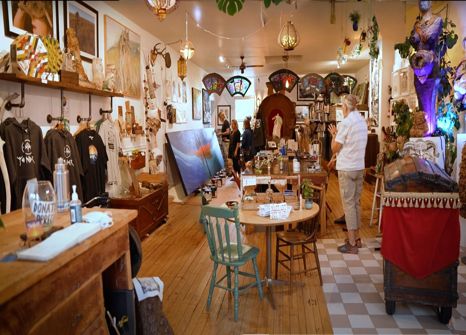 Interior of Yonder's gallery: a warm wood-floored space filled with stained glass lanterns, framed prints, paintings on easels, vintage furniture, and Yonder hoodies on display, with two visitors browsing in the back.