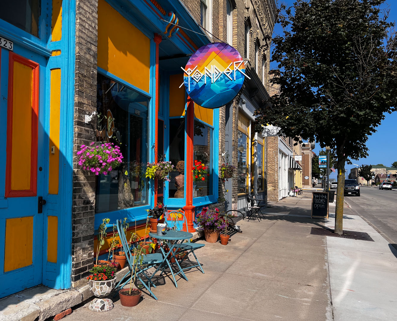 Yonder storefront on a sunny day in downtown Algoma, Wisconsin: a vivid blue-and-orange painted facade in a historic brick building, with a circular Yonder sign featuring a geometric mountain-and-sun mural, hanging flower baskets, and a small turquoise bistro table set out on the sidewalk.